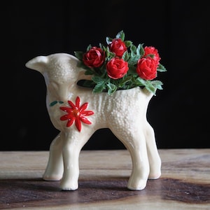 May include: A cream-colored lamb-shaped planter with a red flower design on its side. The planter holds a bouquet of red roses with green leaves. The lamb is standing on four legs.