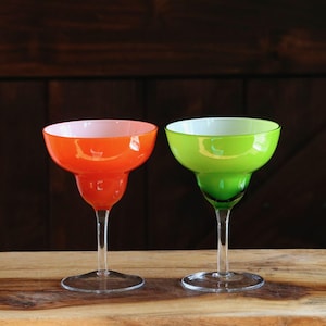 May include: Two margarita glasses with clear stems and bases. One glass is orange with a white interior, and the other is green with a white interior. The glasses are on a wooden surface against a dark brown background.