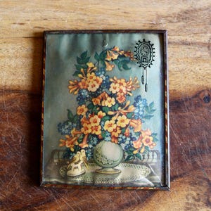 May include: A framed vintage print featuring a still life of flowers in shades of yellow, orange, and blue. A small globe and a decorative shoe are also visible. The frame has a dark brown finish. A clock is drawn on the top right.