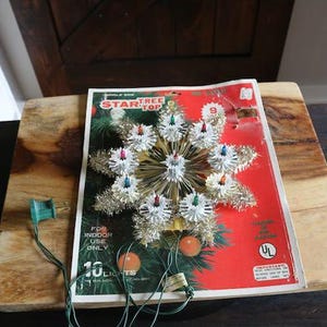 May include: Vintage Christmas tree topper in the shape of a star. The topper has silver tinsel and white lights. The packaging is red and green with the words "Star Tree Top".