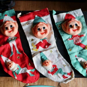 May include: Three vintage Christmas stockings with elf faces and figures. The stockings are red, white, and green, with gold star accents. Each stocking features a smiling elf face and a small elf figure.