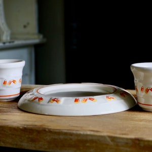 May include: A white ceramic candy corn themed set, including two small cups and a shallow dish. The cups and dish are decorated with orange and yellow candy corn designs and orange trim. The set is displayed on a wooden surface.