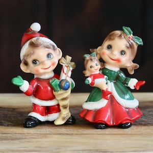 May include: Vintage Christmas figurines of a boy in a Santa suit and a girl in a green and red dress. The boy holds a stocking with a candy cane and a ball. The girl holds a smaller doll. Both have smiling faces.