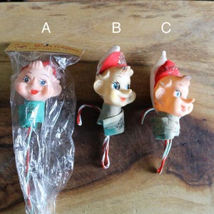 May include: Three vintage Christmas elf ornaments with painted faces, red hats, and green collars. One elf is in a plastic package. The ornaments have candy cane-striped stems and are displayed on a wooden surface.