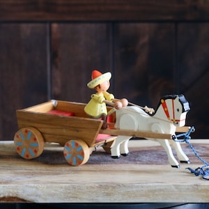 May include: Wooden toy featuring a figure in a yellow coat and red hat driving a cart pulled by a white horse. The cart has red seating and blue and red accents on the wheels. The horse has black and red details.