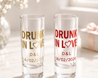 Puede incluir: Dos vasos de chupito de cristal transparente con el texto "DRUNK IN LOVE" y un símbolo de corazón en lugar de la letra O. Un vaso tiene letras doradas y el otro letras rojas. Ambos están personalizados con "D & L" y "14/02/2024".