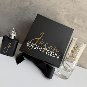 May include: A black hip flask, shot glass, and gift box set. The flask and shot glass have gold lettering. The gift box is black with gold lettering that reads "Jason EIGHTEEN". A black ribbon is tied around the box.