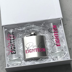 May include: A white gift box containing a silver metal flask and two clear glass shot glasses nestled in white paper shred. The flask and both glasses feature the text Ruby EIGHTEEN in a combination of black script and bold pink block lettering.