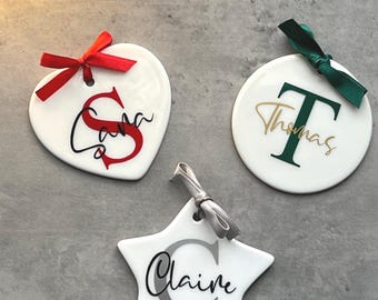 May include: Three white ceramic Christmas ornaments in heart, circle, and star shapes. Each ornament has a name and initial in a different colour. The heart has a red bow, the circle has a green bow, and the star has a silver bow.