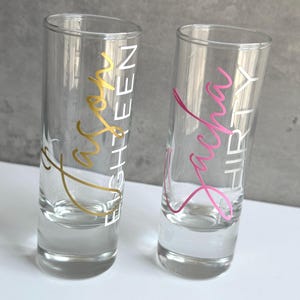May include: Two clear shot glasses with text. One glass has gold text that reads "Jason EIGHTEEN", and the other has pink text that reads "Sasha THIRTY". The glasses are on a white surface with a gray background.