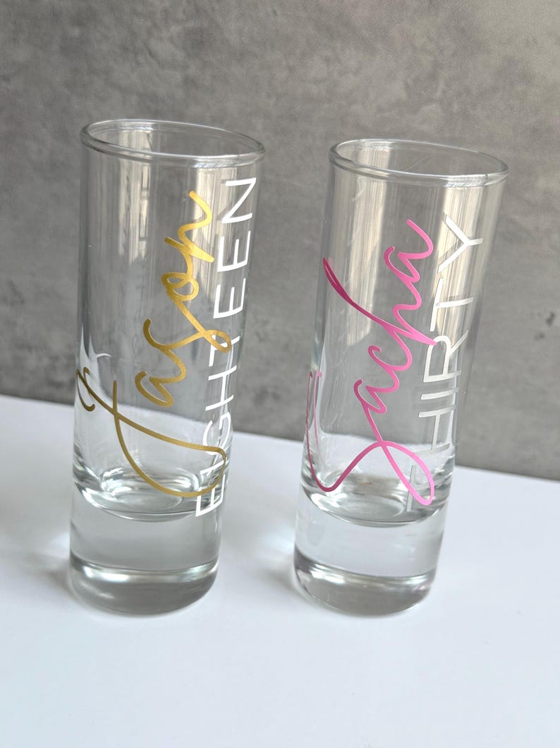 May include: Two clear shot glasses with text. One glass has gold text that reads "Jason EIGHTEEN", and the other has pink text that reads "Sasha THIRTY". The glasses are on a white surface with a gray background.