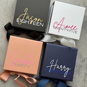 May include: Four square gift boxes in black, white, peach, and navy blue, each with a ribbon. The boxes have names and ages printed on them in various colors: gold, pink, and white. The boxes are on a gray surface.
