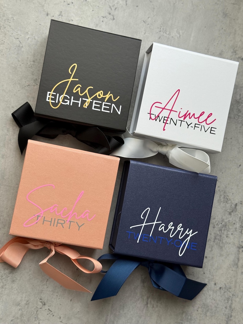 May include: Four square gift boxes in black, white, peach, and navy blue, each with a ribbon. The boxes have names and ages printed on them in various colors: gold, pink, and white. The boxes are on a gray surface.