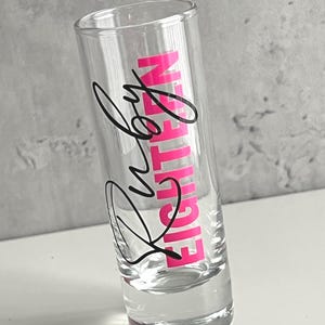 May include: Clear shot glass with the words "Ruby Eighteen" in black and pink lettering. The shot glass is cylindrical and appears to be empty. The background is a light gray.