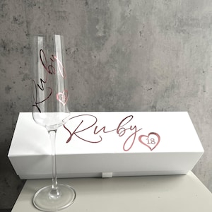 May include: A clear glass champagne flute featuring the name Ruby in rose gold script and a small heart with the number 18. Behind it sits a white rectangular gift box displaying the name Ruby in rose gold script and a heart with the number 18.