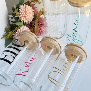 May include: Five clear glass tumblers with bamboo lids and glass straws. Each tumbler features a personalized name in a different color and font style: Elle in black, Katie in pink, Olivia in gold, Jessica in white, and Grace in teal.