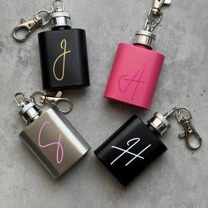 May include: Four small, rectangular flasks with attached keychains. One black flask has a gold "J", a pink flask has a pink "A", a silver flask has a pink "C", and a black flask has a white "H".