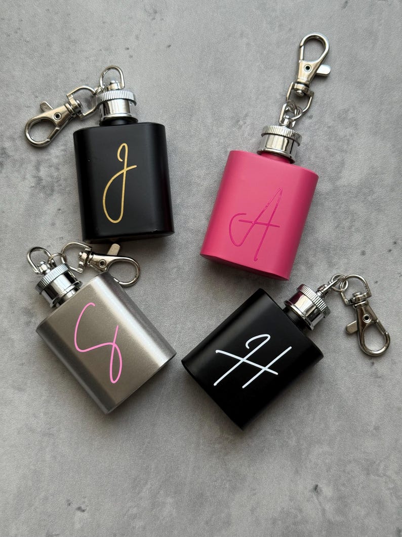 May include: Four small, rectangular flasks with attached keychains. One black flask has a gold "J", a pink flask has a pink "A", a silver flask has a pink "C", and a black flask has a white "H".