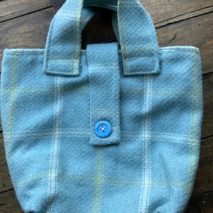Puede incluir: Bolso tote azul claro con estampado de cuadros. El bolso tiene forma rectangular con base redondeada y asa a juego. Un botón azul asegura un cierre de solapa. El bolso está hecho de una tela suave y texturizada.