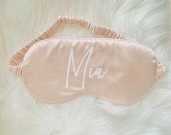 May include: A light peach-colored sleep mask with the name "Mia" printed in white script. The mask has an elastic band for securing around the head. The mask is made of a smooth, satin-like material.