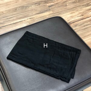 Puede incluir: Un cuadrado de tela negro doblado en tres partes, sobre una superficie de cuero marr&oacute;n. La letra "H" est&aacute; impresa en blanco sobre la tela.