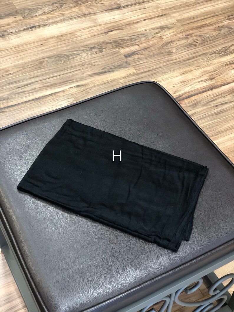 Puede incluir: Un cuadrado de tela negro doblado en tres partes, sobre una superficie de cuero marr&oacute;n. La letra "H" est&aacute; impresa en blanco sobre la tela.