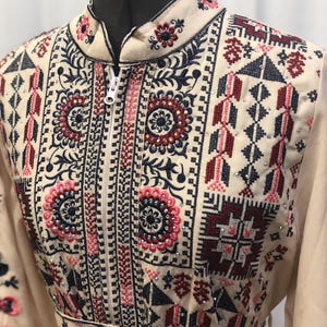 May include: A cream-colored long-sleeved dress with a zipper closure and intricate embroidery in shades of red, pink, blue, and black. The embroidery features geometric patterns and floral motifs, accented with sparkling rhinestones.