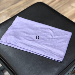 Puede incluir: Una diadema de tela de color lila claro, doblada y colocada sobre una superficie de cuero negro. La letra "D" es visible en la diadema.