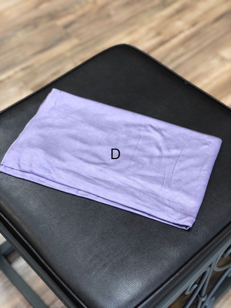 Puede incluir: Una diadema de tela de color lila claro, doblada y colocada sobre una superficie de cuero negro. La letra "D" es visible en la diadema.