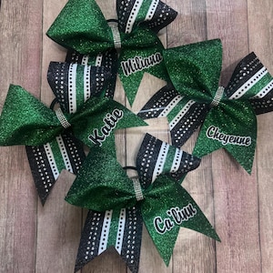 May include: A collection of green glitter cheerleading bows with black and white accents. Each bow features a rhinestone center and a name in white script. The bows are arranged on a wooden surface.