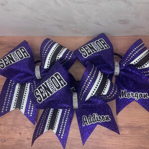 May include: Purple glitter cheerleading bows with black and white accents and the word "Senior 2026". Some bows have names like "Addison" and "Morgan". Each bow has a rhinestone center.