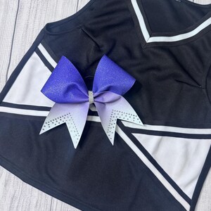 May include: A black and white cheerleading uniform top with a V-neck and a large purple and white bow. The bow has a rhinestone center and rhinestone accents on the tips. The top has white stripes and geometric designs.