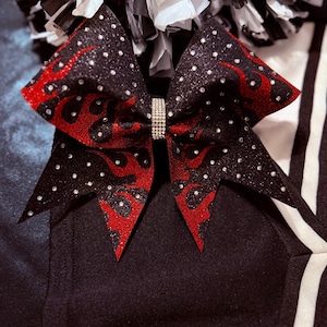 Può includere: Un fiocco da cheerleader nero con accenti di fiamme rosse e strass scintillanti. Il fiocco presenta un centro di strass ed è ambientato su uno sfondo a righe bianche e nere. Accessorio da cheerleader.