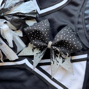 May include: A black glitter hair bow featuring silver flame patterns on the tails and small clear rhinestones across the top loops, centered with a band of silver rhinestones, resting on a black and white cheerleading uniform and pom-poms.
