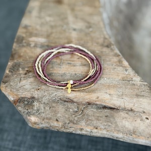 May include: A set of four stacked beaded bracelets featuring small round beads in shades of plum, metallic gold, and cream. The bracelets are accented with a single small white bead and a gold-toned metal charm shaped like a leaf or feather.