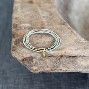 Könnte beinhalten: Ein mehrreihiges Perlenarmband mit abwechselnden Reihen aus hellblauen und goldfarbenen Saatperlen. Das Armband enthält eine kleine weiße Perle und einen hängenden goldfarbenen Anhänger in Form eines stilisierten Blattes oder Auges.
