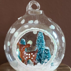 May include: A glass Christmas ornament with white polka dots and a snowy scene inside. A small brown deer figurine stands in the snow with a blue pine tree behind it.