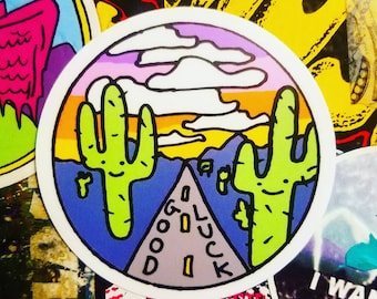 May include: A round sticker with a desert scene. Two green cacti flank a gray road with the words "GOOD LUCK." The background features a sunset with orange, yellow, and purple hues, with white clouds above. The sticker is surrounded by other stickers.