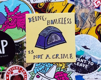 Könnte beinhalten: Ein gelber quadratischer Aufkleber mit dem Text "BEING HOMELESS IS NOT A CRIME" und einer Zeichnung eines Zeltes. Der Aufkleber ist von anderen Aufklebern mit verschiedenen Designs und Texten umgeben.