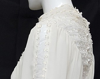 Puede incluir: Vestido color marfil con mangas largas y falda fluida. Las mangas y el escote están adornados con delicados adornos. Se ve una pulsera negra en la muñeca del maniquí. El vestido es una prenda formal.