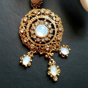 Puede incluir: Collar con colgante dorado con diseño de filigrana. El colgante presenta una gran piedra central opalescente rodeada de piedras claras más pequeñas. Tres piedras opalescentes en forma de lágrima cuelgan debajo. La cadena también es dorada.