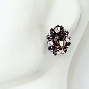 May include: An oval-shaped stud earring with a central brown gemstone, surrounded by smaller clear and dark gemstones. The earring is set in a dark metal and has a classic, elegant design.