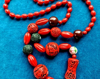 May include: A red beaded necklace with carved wooden beads in various colours, including green, brown, and white. The necklace features intricate floral designs and patterns.