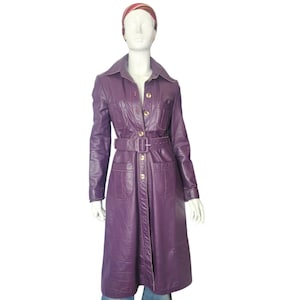 May include: A long, purple leather coat with a belted waist and gold-coloured buttons. The coat has a collared neckline and two front pockets. The mannequin is also wearing a patterned headband and blue jeans.