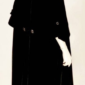 Puede incluir: Un abrigo de terciopelo negro con cuello de capa y mangas largas. El abrigo tiene una cintura ajustada y una falda acampanada. El abrigo lo lleva puesto un maniqu&iacute; con un turbante blanco.