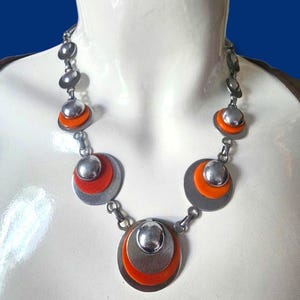 Puede incluir: Un collar plateado con colgantes circulares. Cada colgante presenta un diseño en capas con un disco plateado, un acento naranja vibrante y una cúpula plateada más pequeña. Se muestra en un maniquí blanco.