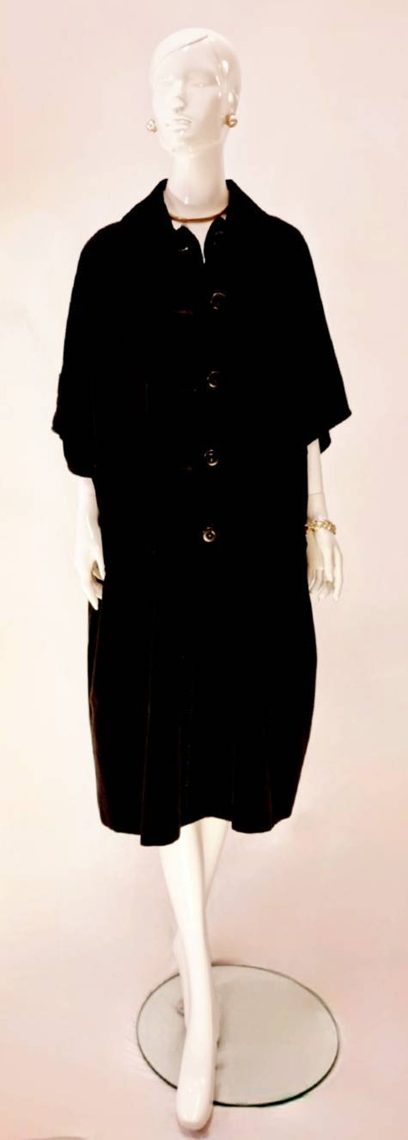 Puede incluir: Un vestido de terciopelo negro con botones en la parte delantera y mangas cortas. El vestido est&aacute; en un maniqu&iacute; blanco.