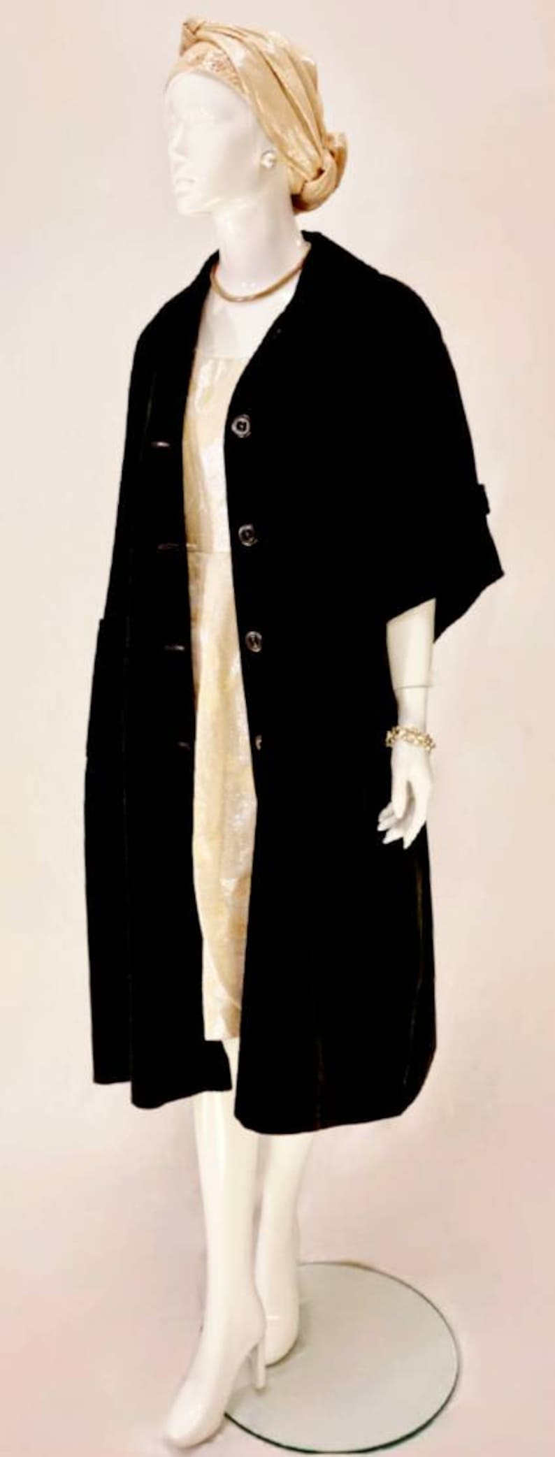 Puede incluir: Un abrigo de terciopelo negro con cuello alto y tres botones. El abrigo tiene mangas cortas y un corte holgado. El maniqu&iacute; lleva un collar de oro y una diadema de oro.