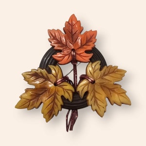 Peut inclure: Une broche vintage représentant trois feuilles d'érable sculptées dans des tons d'orange, de jaune et de brun. Les feuilles sont disposées autour d'une base circulaire brun foncé.