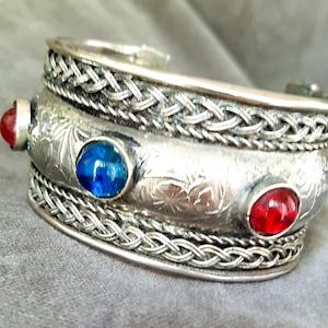 Peut inclure: Bracelet manchette argenté avec des motifs tressés et floraux. Il comporte deux pierres rouges et une bleue. Le bracelet a une surface détaillée et texturée. Style vintage.